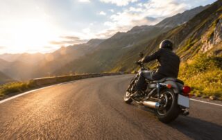 Ein Motorradfahrer fährt auf einer kurvenreichen Straße in den Alpen, während die Sonne hinter den Bergen untergeht.